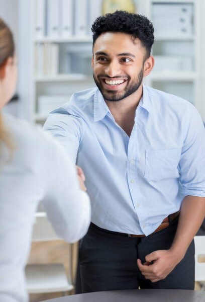 An attractive young man smiles broadly as he stands and leans across a table to shake hands with his unrecognizable new female manager.  She has just offered him a job on the spot.